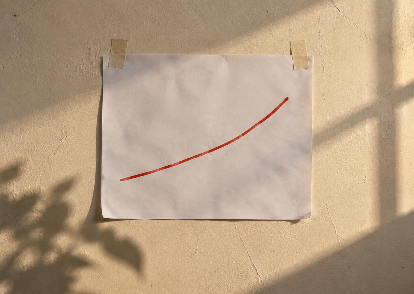 Ett papper med en handritad röd stigande kurva tejpflat på en varm kremvägg i eftermiddagsljus.